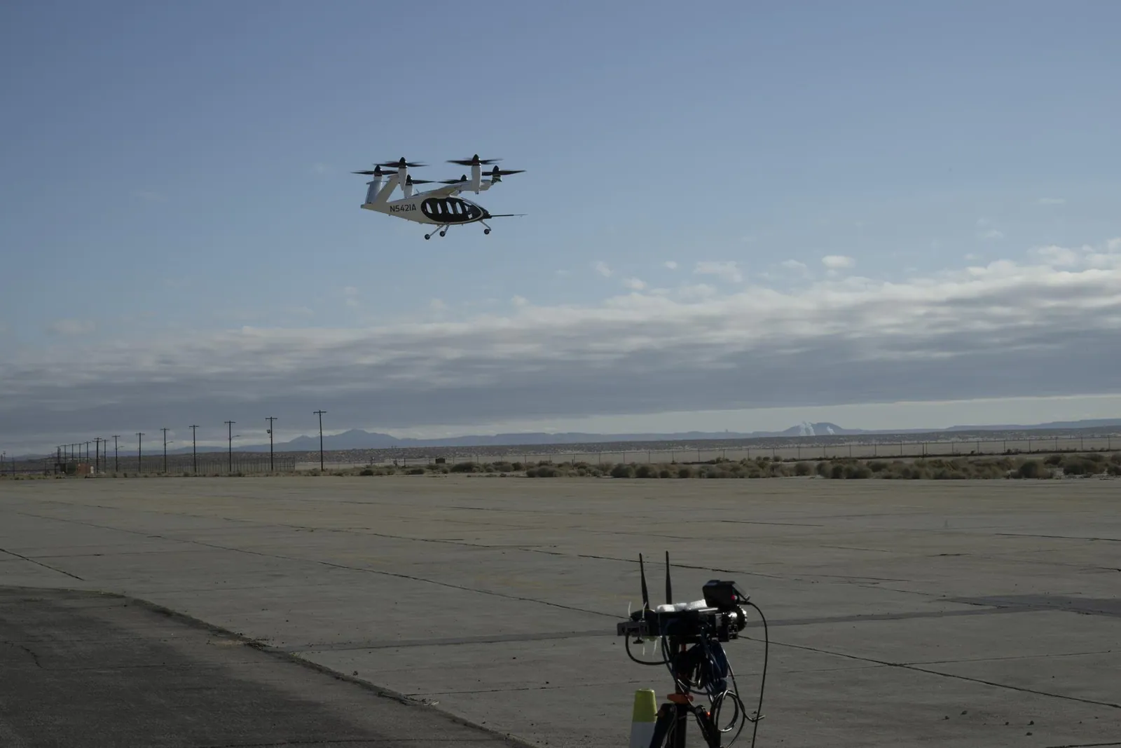 An electric air taxi hovers over an airfield, with a sensor on a tripod in the foreground.