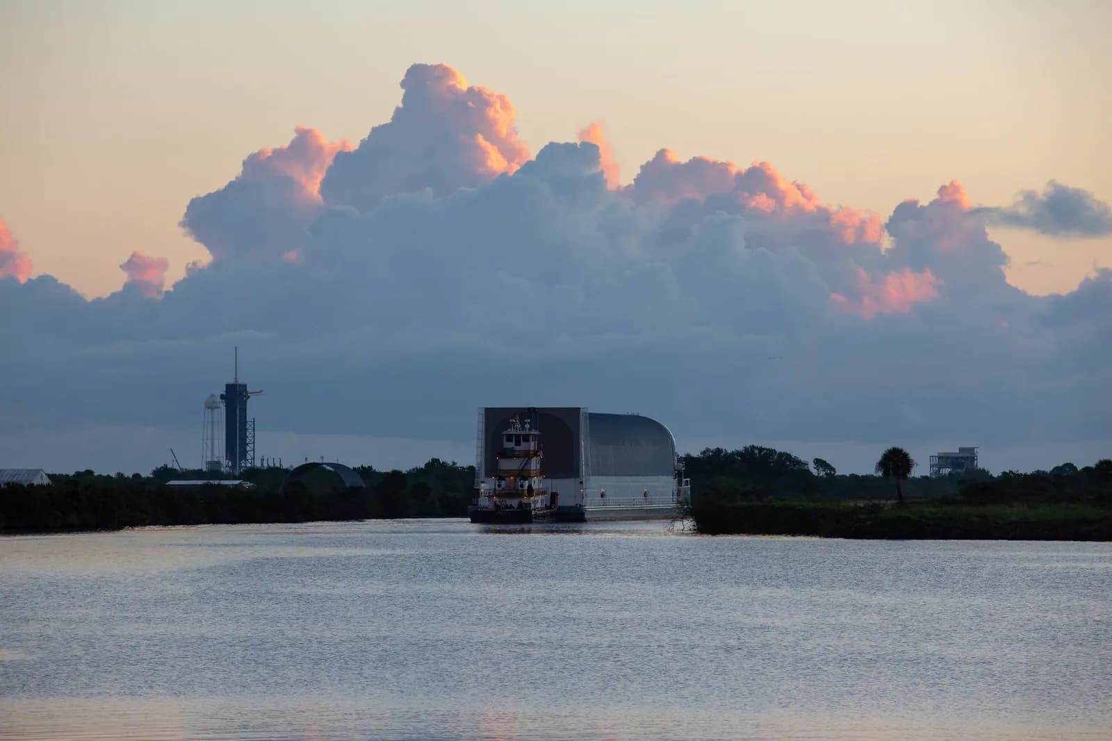 A barge carrying a full-size rocket core stage crosses calm water at dusk.