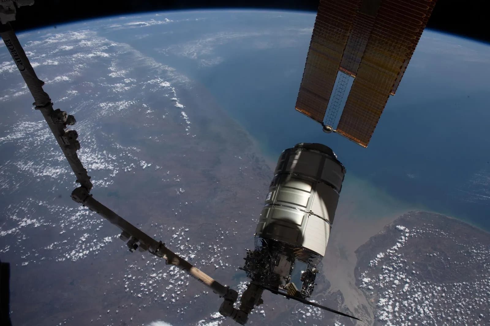 A space station robotic arm holds a cylindrical cargo spacecraft above Earth's coastline.