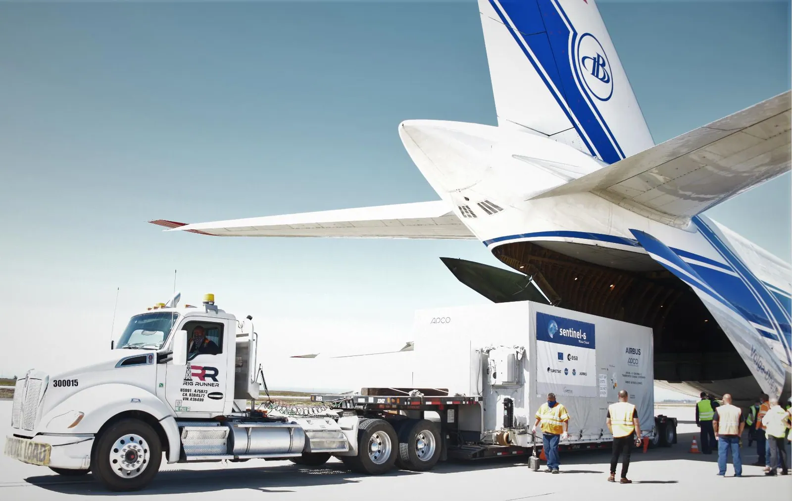 A satellite in its shipping container is driven away from a large cargo aircraft on an airfield.
