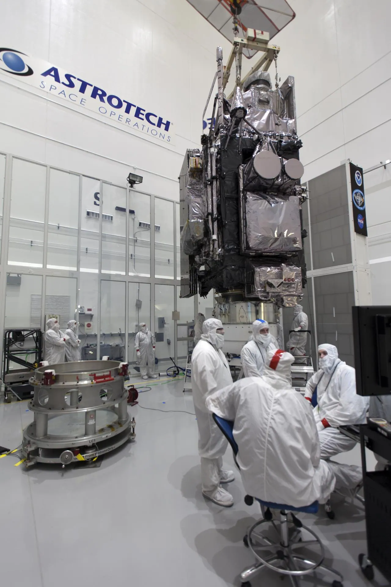 Technicians in cleanroom suits work beside a large satellite in a clean room.